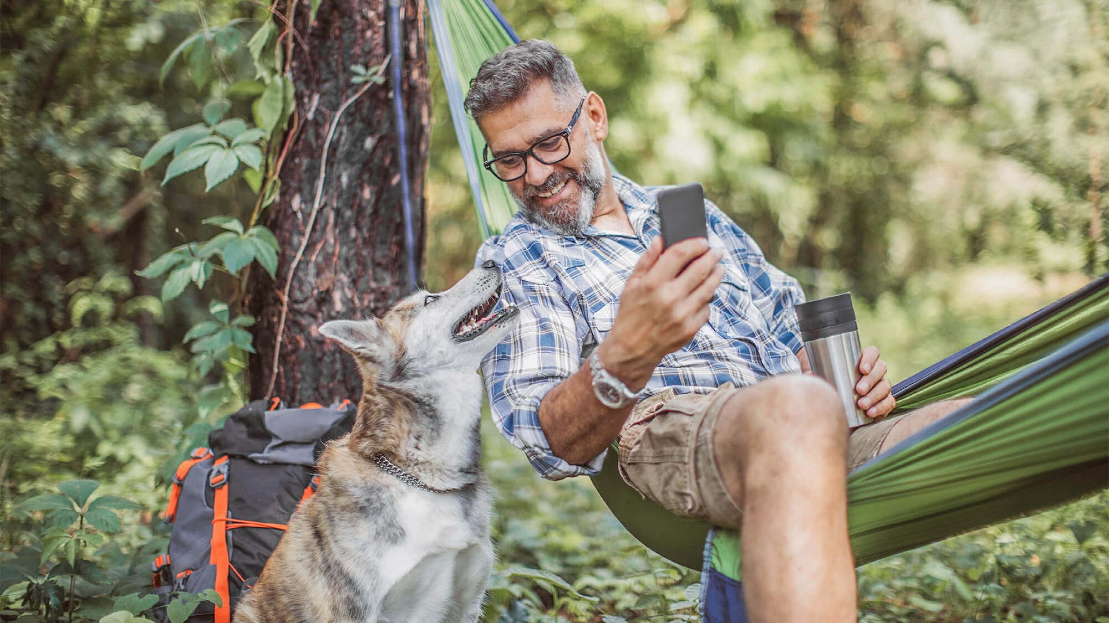 Zufriedener älterer Mann in Hängematte mit Handy und Thermobecher, neben ihm sitzt sein Hund und Blickt ihn an
